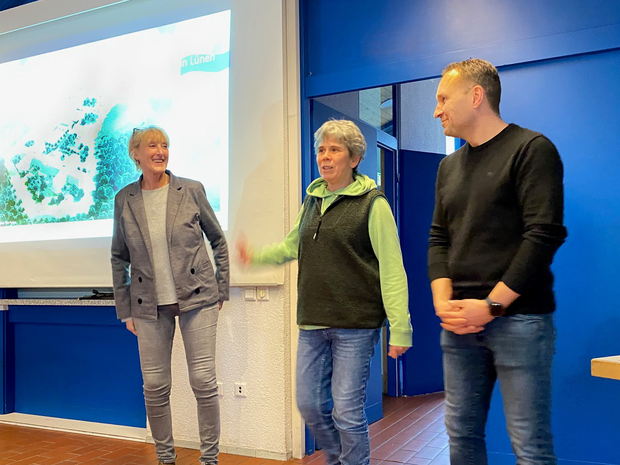 Zwei Frauen und ein Mann stehen lächelnd vor einer beleuchteten Leinwand. Die Frau in der Mitte stellt die anderen vor. (Bild: LWL/SchuFi)