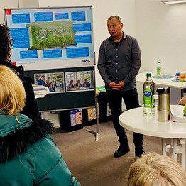 Ein Mann steht vor einem Plakat und spricht mit einer Gruppe von Menschen. (Bild: LWL/SchuFi)