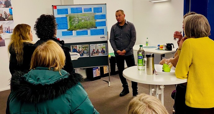 Ein Mann steht vor einem Plakat und spricht zu einer Gruppe von Menschen. (Bild: LWL-SchuFi)