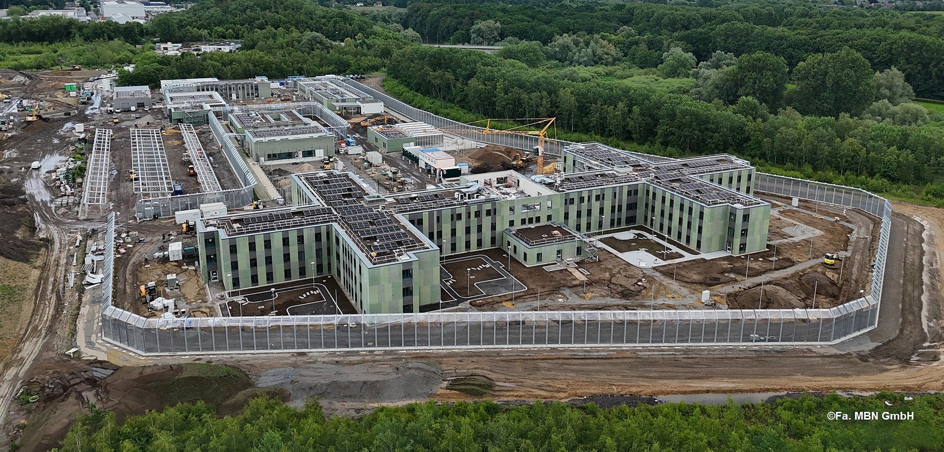 Luftaufnahme mehrerer grün verklinkerter Gebäude, umgeben von einer Zaunanlage und Wald (Bild: Fa. MBN GmbH)